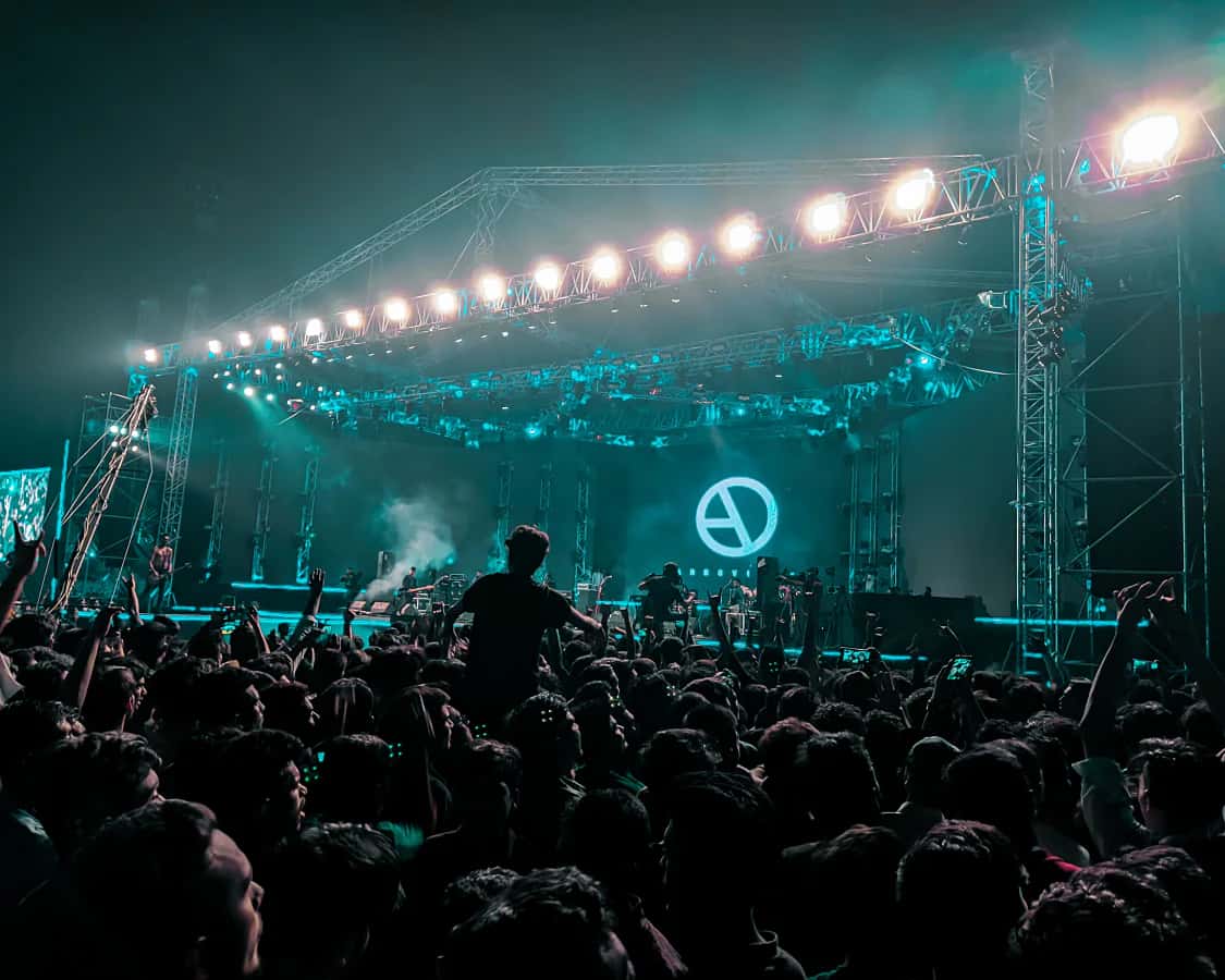 A packed fest crowd with hands up in front of a teal-lit stage