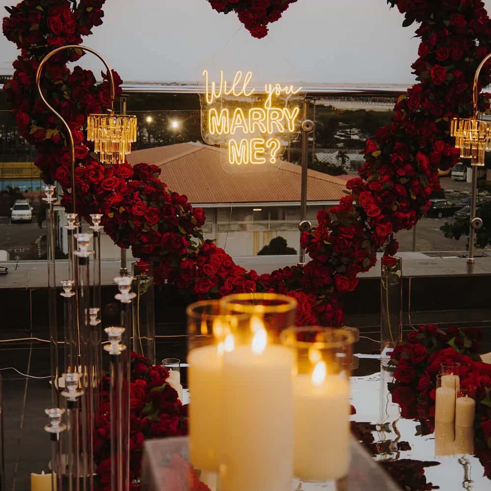 A red-rose heart arch with a “Will you marry me?” neon over lit candles