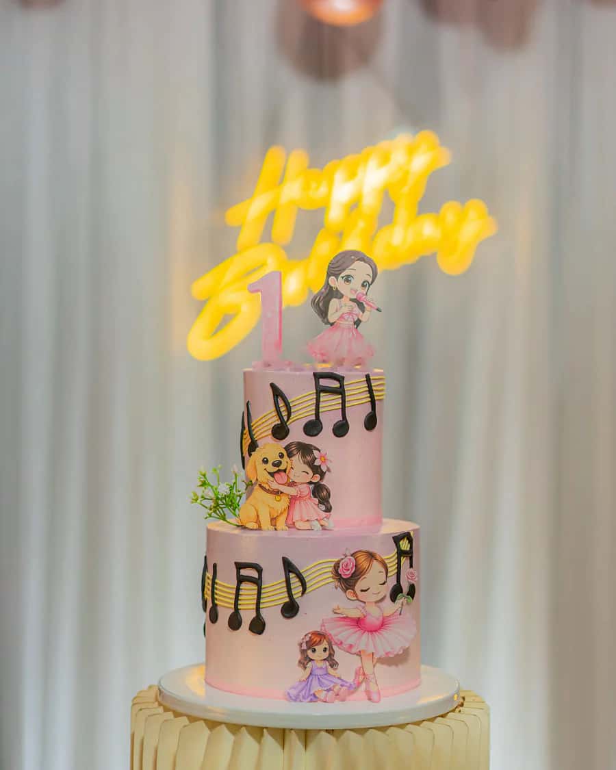 A two-tier pink birthday cake with music notes under a glowing Happy Birthday neon